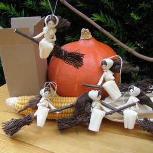 May include: Handmade corn husk witch ornaments on a broomstick, with a pumpkin and corn. The witches have brown hair and are wearing white dresses and bonnets. A cardboard box is in the background. The scene is set on a wooden surface.