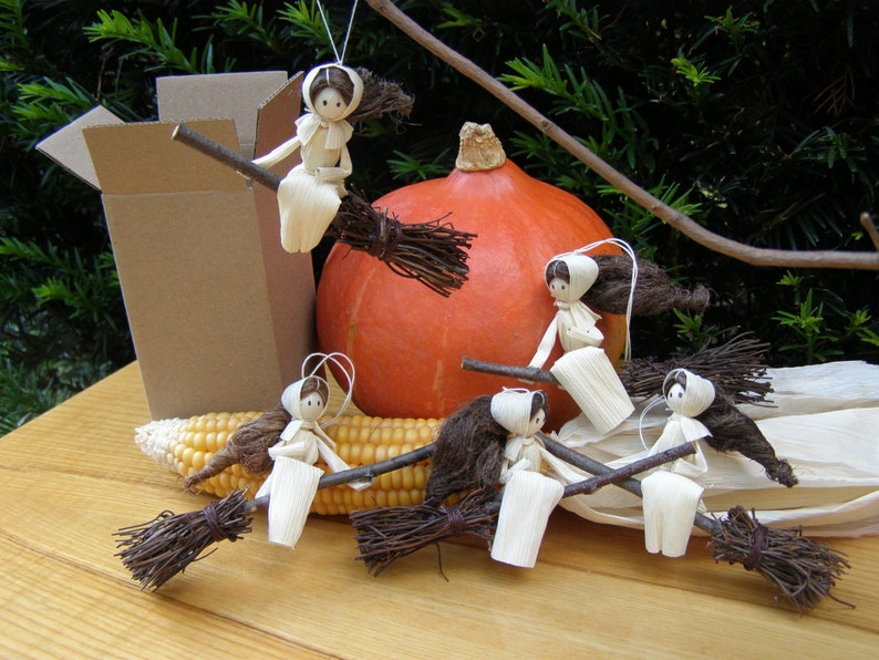 May include: Handmade corn husk witch ornaments on a broomstick, with a pumpkin and corn. The witches have brown hair and are wearing white dresses and bonnets. A cardboard box is in the background. The scene is set on a wooden surface.