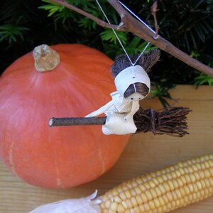 May include: A decorative Halloween-themed display featuring a large orange pumpkin, a miniature witch figurine riding a broom, and a cob of corn. The witch is crafted from natural materials. The scene evokes autumn and harvest themes.