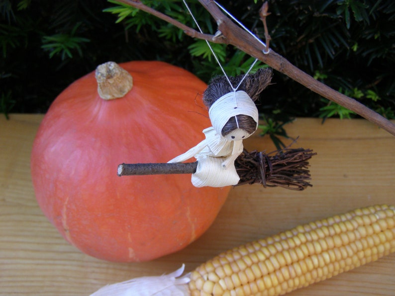 May include: A decorative Halloween-themed display featuring a large orange pumpkin, a miniature witch figurine riding a broom, and a cob of corn. The witch is crafted from natural materials. The scene evokes autumn and harvest themes.