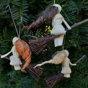 May include: Handmade witch ornaments, each crafted with natural materials. The witches have straw-like hair in various colors, wearing white dresses, and riding on twig brooms. These whimsical decorations are suspended by white strings.