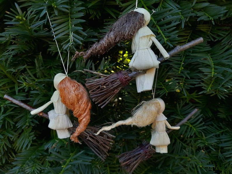 May include: Handmade witch ornaments, each crafted with natural materials. The witches have straw-like hair in various colors, wearing white dresses, and riding on twig brooms. These whimsical decorations are suspended by white strings.