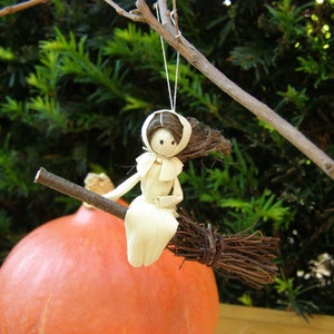 May include: A handmade corn husk witch doll riding a twig broom. The doll has a white dress and a brown hair. The broom is made of twigs and is attached to a branch. The background includes a large orange pumpkin and green foliage.
