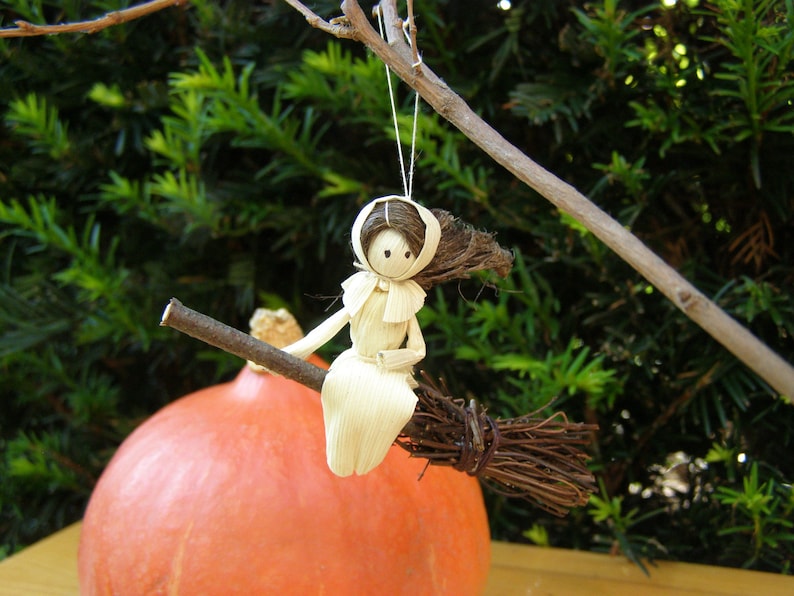 May include: A handmade corn husk witch doll riding a twig broom. The doll has a white dress and a brown hair. The broom is made of twigs and is attached to a branch. The background includes a large orange pumpkin and green foliage.