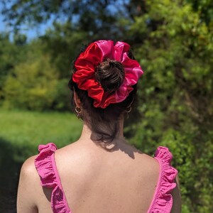 Puede incluir: Una mujer con cabello oscuro recogido en un mo&ntilde;o, adornado con una gran scrunchie roja y rosa. Lleva un vestido rosa con tirantes con volantes. El fondo presenta una valla de madera, hierba verde y &aacute;rboles bajo un cielo azul.