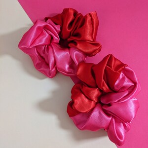 Puede incluir: Dos coleteros de sat&eacute;n en tonos rojos y rosas. Los coleteros est&aacute;n colocados sobre un fondo rosa y blanquecino. La tela tiene una textura brillante y suave.