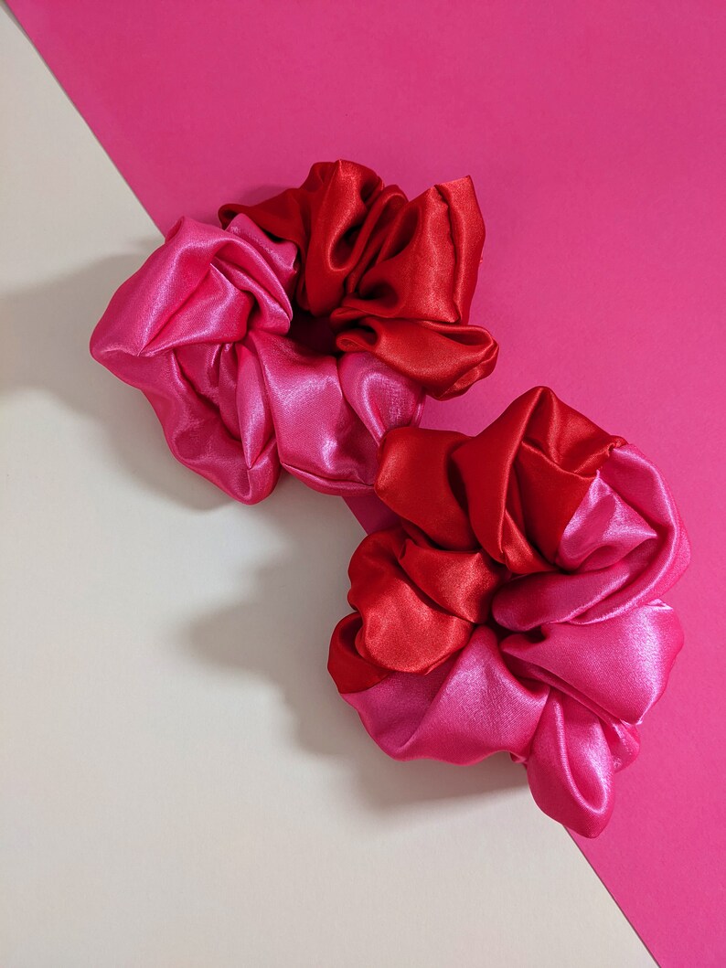 Puede incluir: Dos coleteros de sat&eacute;n en tonos rojos y rosas. Los coleteros est&aacute;n colocados sobre un fondo rosa y blanquecino. La tela tiene una textura brillante y suave.