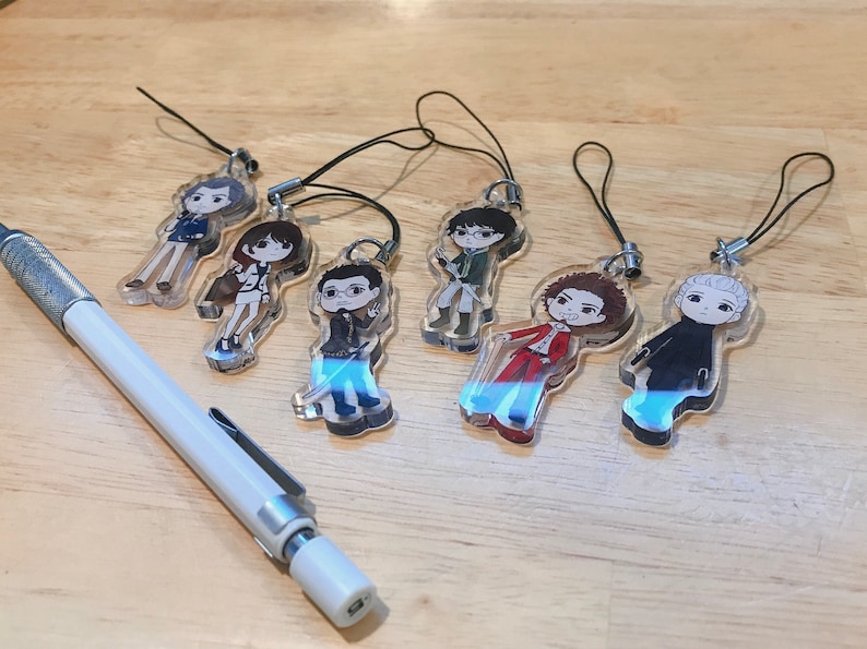 May include: Seven colorful character charms on clear acrylic keychains with black cords. The characters are drawn in a cartoon style and feature different hairstyles, clothing, and expressions.