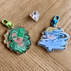 May include: Two clear acrylic keychains with colourful illustrations. The keychain on the left features a green and white illustration of a person with a card over their eye. The keychain on the right features a blue and white illustration of a person with a cloud over their eye. Both keychains have metal rings.