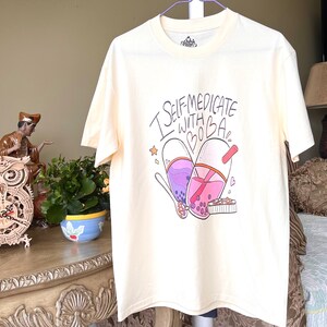 May include: A white t-shirt featuring a graphic of two boba tea cups shaped like pills. The text "I self-medicate with boba" is written above the cups.