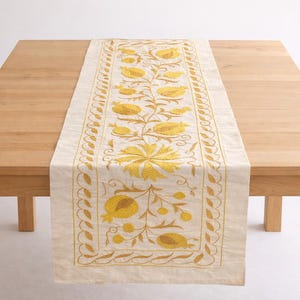 Könnte beinhalten: Ein rechteckiger, cremefarbener Tischläufer mit gelbem Blumendesign. Das florale Muster zeigt stilisierte Blumen und Blätter. Der Tischläufer liegt auf einem hellen Holztisch.