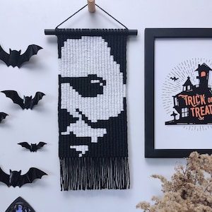 May include: Black and white macrame wall hanging with a skull design. The hanging is framed with a black wooden dowel and has a fringe at the bottom. The hanging is displayed on a white wall with other Halloween decorations.