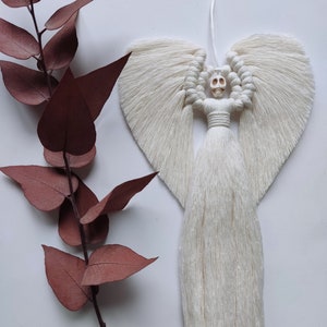 May include: White macrame angel with a skull head and long fringe. The angel is hanging from a white string.