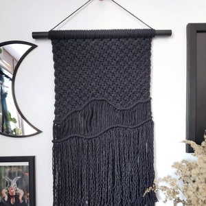 Könnte beinhalten: Schwarze Makramee-Wandbehang mit wellenförmigem Muster und langen Fransen. Der Wandbehang ist aus dickem schwarzem Seil gefertigt und an einer schwarzen Holzleiste befestigt.