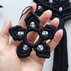 May include: Black macrame wall hanging with a starburst design featuring white skull beads. The wall hanging has a long black tassel.