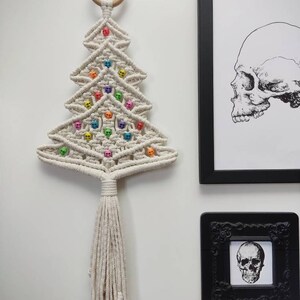 K&ouml;nnte beinhalten: Ein wei&szlig;er Makramee-Weihnachtsbaum-Wandbehang mit einem Holzring oben. Der Baum ist mit bunten Totenkopfperlen verziert.