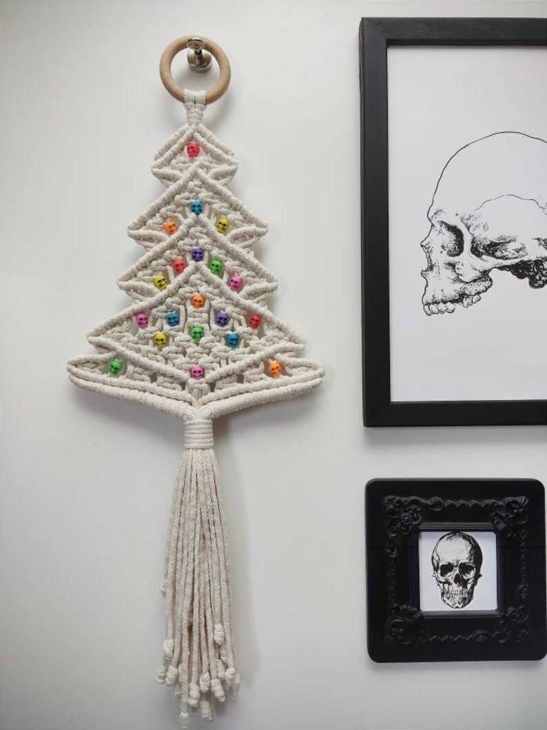 K&ouml;nnte beinhalten: Ein wei&szlig;er Makramee-Weihnachtsbaum-Wandbehang mit einem Holzring oben. Der Baum ist mit bunten Totenkopfperlen verziert.