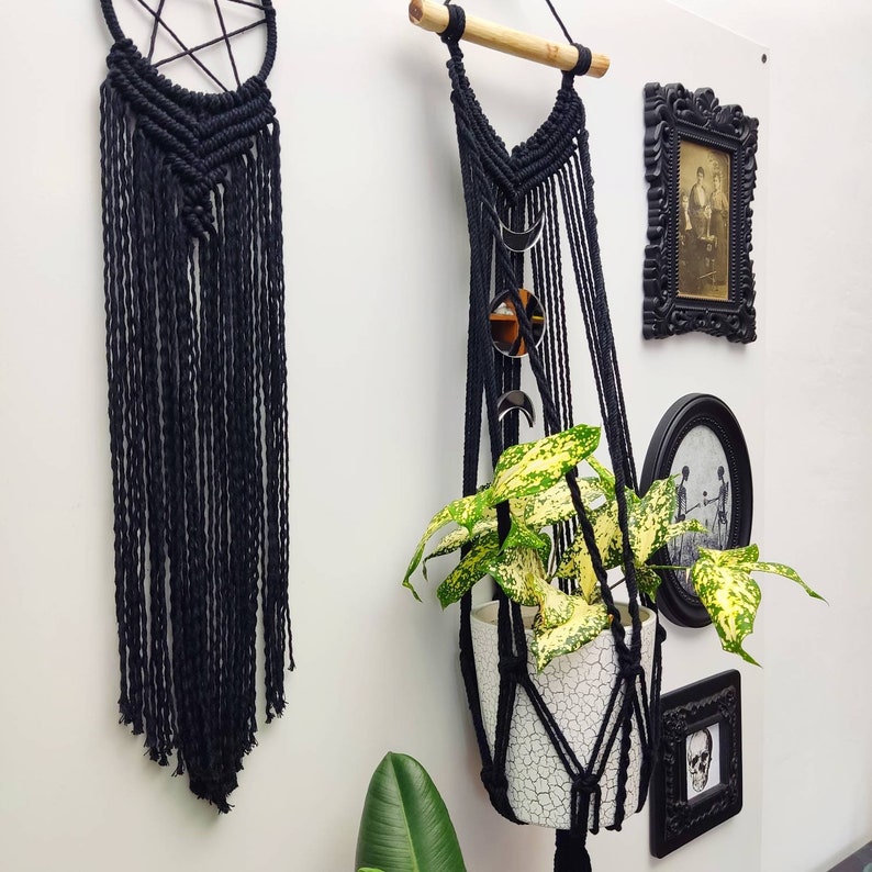 Op de afbeelding: Een zwarte macrame plantenhanger met een witte pot en een groene plant met bonte bladeren. De hanger is versierd met zilveren halve manen en een ronde spiegel.
