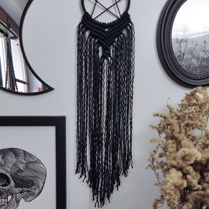 Op de afbeelding: Zwarte macram&eacute; droomvanger met een pentagram-ontwerp en lange franjes. De droomvanger hangt aan een witte muur met een zwart-witte schedelprint en een zwarte spiegel op de achtergrond.