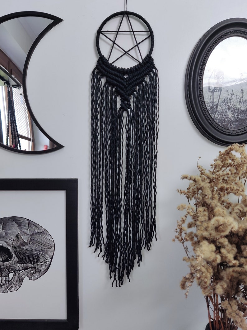 Op de afbeelding: Zwarte macram&eacute; droomvanger met een pentagram-ontwerp en lange franjes. De droomvanger hangt aan een witte muur met een zwart-witte schedelprint en een zwarte spiegel op de achtergrond.