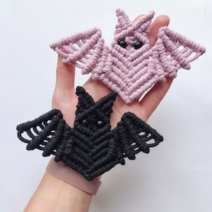 May include: Two handmade macrame bats, one black and one pink, with black bead eyes. The bats are hanging from a person's hand.