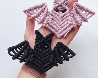 May include: Two handmade macrame bats, one black and one pink, with black bead eyes. The bats are hanging from a person's hand.