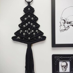 K&ouml;nnte beinhalten: Schwarzer Makramee-Wandbehang in Form eines Weihnachtsbaums mit wei&szlig;en Totenkopfperlen. Der Baum hat eine lange schwarze Quaste.
