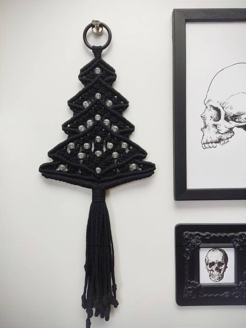K&ouml;nnte beinhalten: Schwarzer Makramee-Wandbehang in Form eines Weihnachtsbaums mit wei&szlig;en Totenkopfperlen. Der Baum hat eine lange schwarze Quaste.