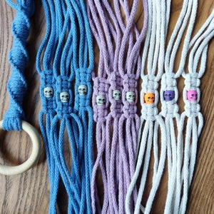 May include: Three macrame plant hangers with skull beads. The hangers are made of white, purple, and blue cord. The skull beads are white, purple, and orange.