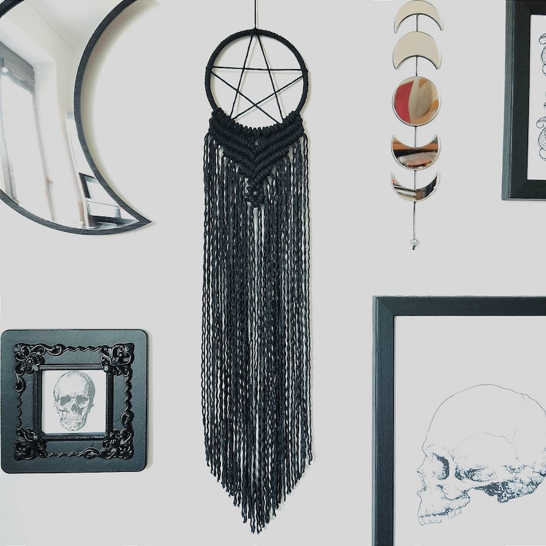 Op de afbeelding: Zwarte macram&eacute; droomvanger met een pentagram-ontwerp en lange franjes die aan een witte muur hangen.