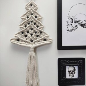 K&ouml;nnte beinhalten: Ein wei&szlig;er Makramee-Weihnachtsbaum-Wandbehang mit schwarzen Perlen. Der Baum hat einen Quasten am unteren Ende und h&auml;ngt an einem schwarzen Metallring.