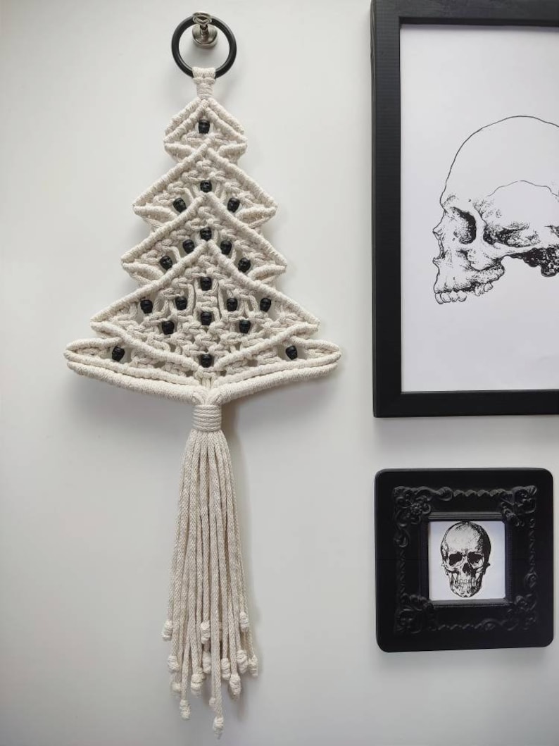 K&ouml;nnte beinhalten: Ein wei&szlig;er Makramee-Weihnachtsbaum-Wandbehang mit schwarzen Perlen. Der Baum hat einen Quasten am unteren Ende und h&auml;ngt an einem schwarzen Metallring.
