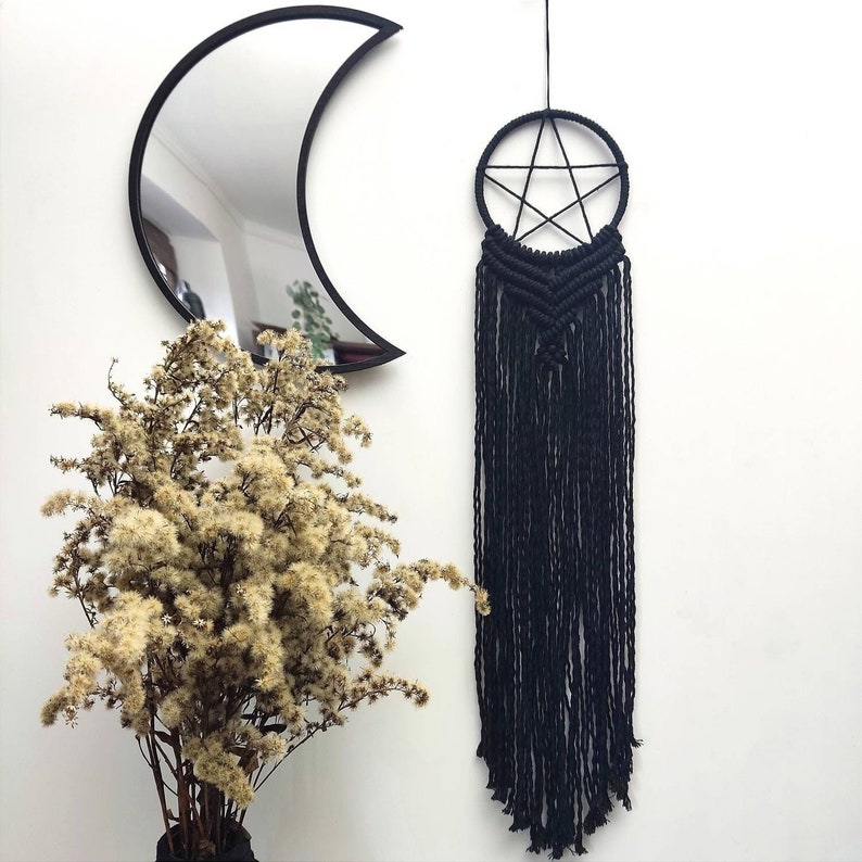 Op de afbeelding: Zwarte macram&eacute; wandhanger met een pentagram-ontwerp en lange franjes. De hanger hangt tegen een witte muur met een zwarte halve maanspiegel en gedroogde bloemen in een vaas.
