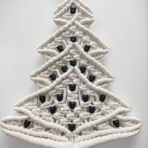 K&ouml;nnte beinhalten: Ein wei&szlig;er Makramee-Weihnachtsbaum-Wandbehang mit schwarzen Totenkopfperlen. Der Baum ist mit einer Knotentechnik gefertigt und hat unten einen Quasten.