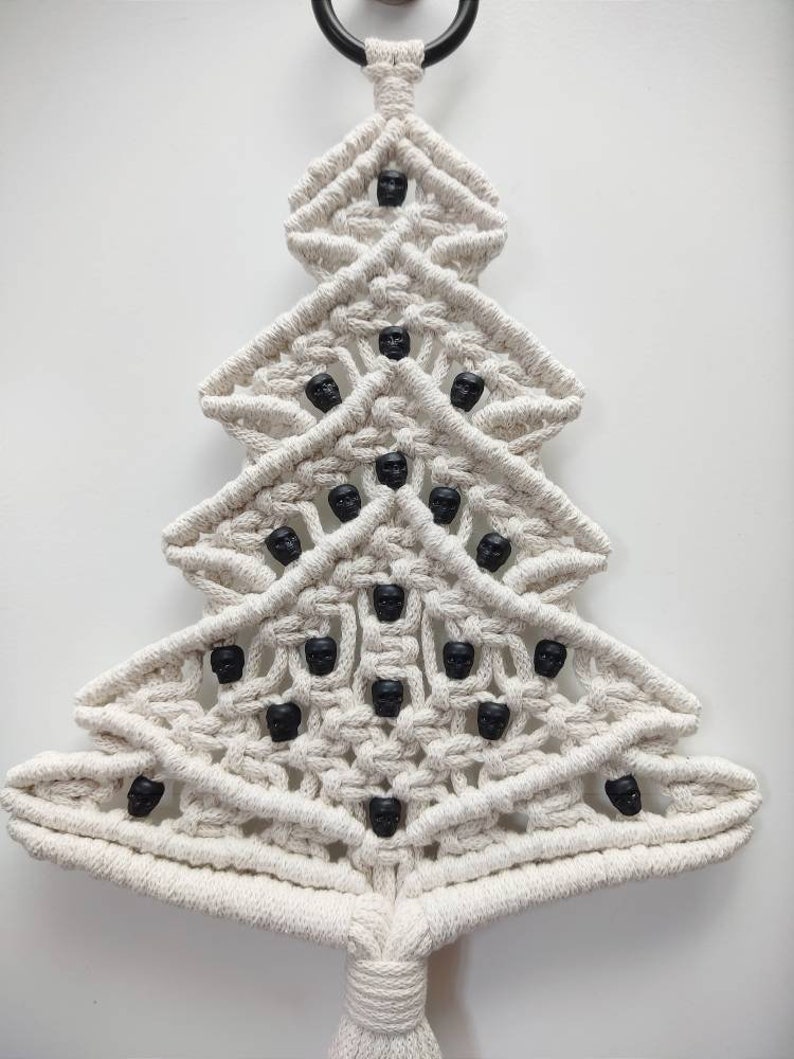 K&ouml;nnte beinhalten: Ein wei&szlig;er Makramee-Weihnachtsbaum-Wandbehang mit schwarzen Totenkopfperlen. Der Baum ist mit einer Knotentechnik gefertigt und hat unten einen Quasten.