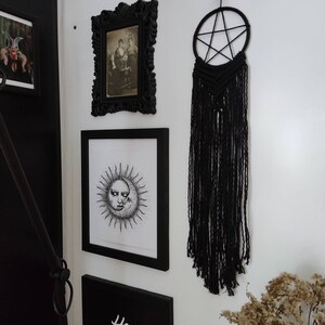 Op de afbeelding: Zwart-witte illustratie van een zon en maan met een menselijk gezicht, ingelijst in een zwart frame, hangend aan een witte muur met een zwarte macram&eacute; droomvanger met een stervormig ontwerp.