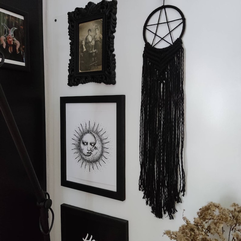 Op de afbeelding: Zwart-witte illustratie van een zon en maan met een menselijk gezicht, ingelijst in een zwart frame, hangend aan een witte muur met een zwarte macram&eacute; droomvanger met een stervormig ontwerp.