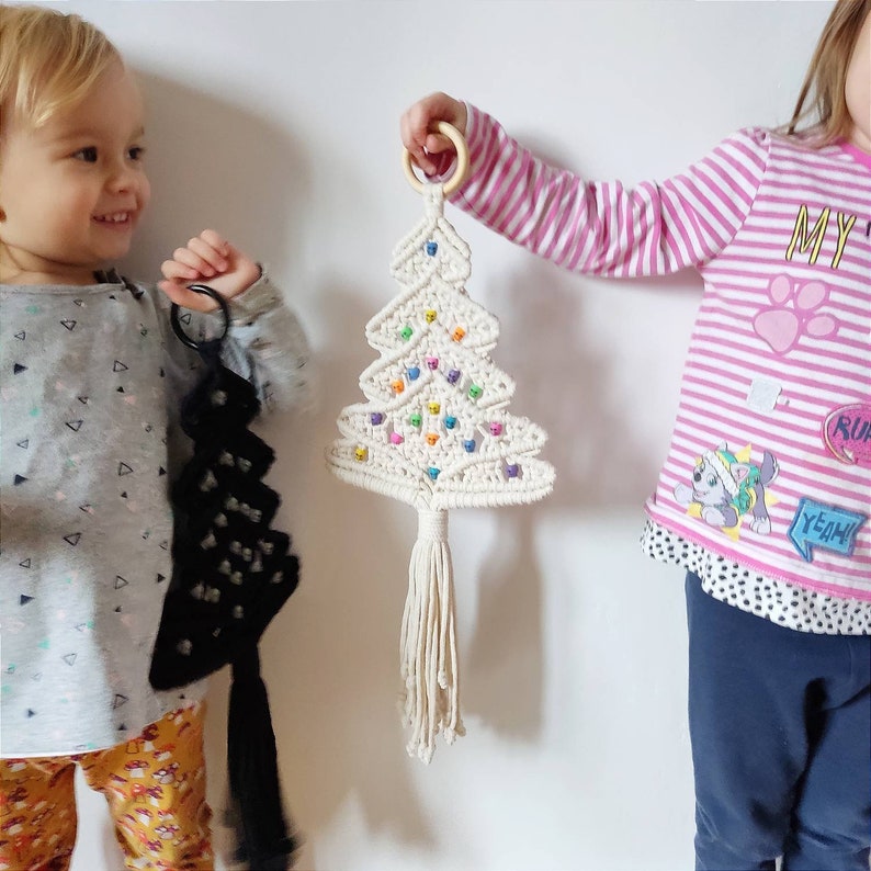 K&ouml;nnte beinhalten: Zwei Kinder, eines h&auml;lt einen wei&szlig;en Makramee-Weihnachtsbaum mit bunten Perlen, das andere h&auml;lt einen schwarzen Makramee-Baum.