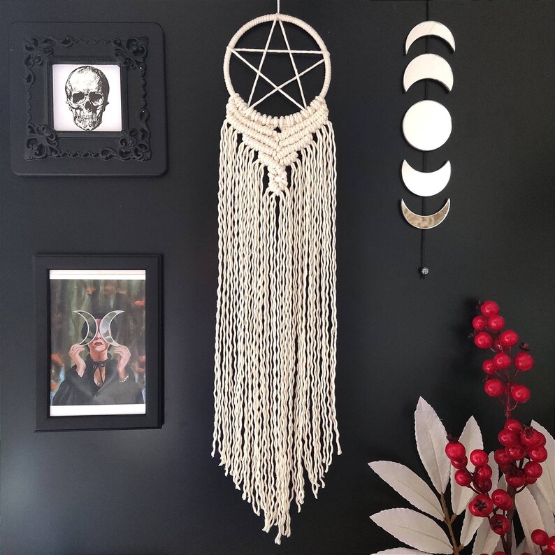 Op de afbeelding: Een witte macram&eacute; droomvanger met een vijfpuntige ster in het midden, hangend aan een zwarte muur met zilveren halve maan wanddecoratie.