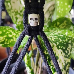 Może przedstawiać: Czarny makramowy wieszak na rośliny z małą, kremową koralikiem w kształcie czaszki pośrodku. Wieszak znajduje się przed zieloną rośliną z białymi plamami, tworząc kontrast.