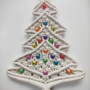 K&ouml;nnte beinhalten: Ein wei&szlig;er Makramee-Weihnachtsbaum-Wandbehang mit einem Holzring oben und einem Quasten unten. Der Baum ist mit bunten Totenkopfperlen verziert.
