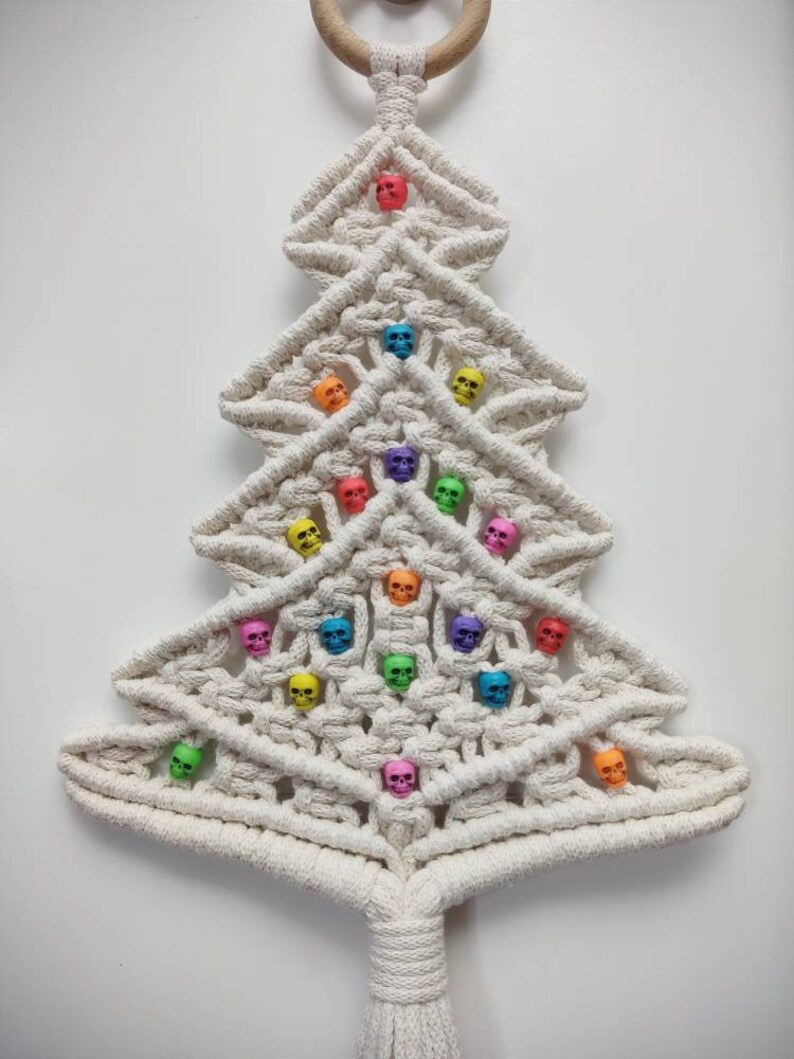 K&ouml;nnte beinhalten: Ein wei&szlig;er Makramee-Weihnachtsbaum-Wandbehang mit einem Holzring oben und einem Quasten unten. Der Baum ist mit bunten Totenkopfperlen verziert.