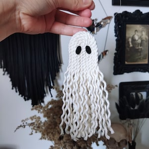 Könnte beinhalten: Weißer Geister-Anhänger aus Garn mit schwarzen Augen. Der Geist hängt an einer weißen Schnur.