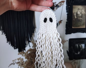 May include: White yarn ghost ornament with black eyes. The ghost is hanging from a white string.