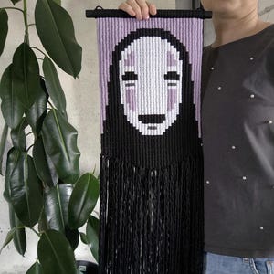 以下が含まれることがあります： 黒と白の織られた壁掛けで、紫と白の顔に笑顔が描かれています。顔は黒いフリンジで囲まれています。