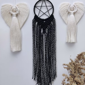 Op de afbeelding: Een zwarte macram&eacute; droomvanger met een vijfpuntige stervorm hangt aan een witte muur. Twee witte macram&eacute; engelen met schedelgezichten hangen aan weerszijden van de droomvanger.