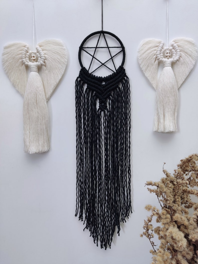 Op de afbeelding: Een zwarte macram&eacute; droomvanger met een vijfpuntige stervorm hangt aan een witte muur. Twee witte macram&eacute; engelen met schedelgezichten hangen aan weerszijden van de droomvanger.