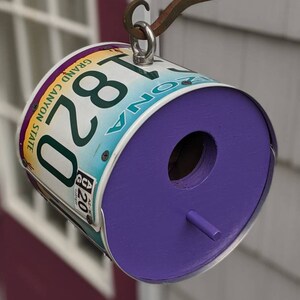 May include: A purple birdhouse made from a repurposed Arizona license plate with the number 1820. The birdhouse is hanging from a metal hook.