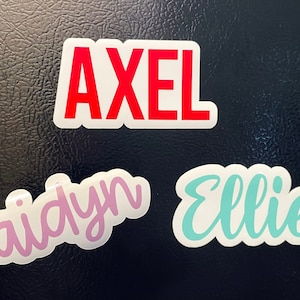 May include: Three name stickers: "Axel" in red block letters, "Jaidyn" in pink script, and "Ellie" in turquoise script. All names are on white backgrounds. The stickers are displayed on a textured, dark surface.