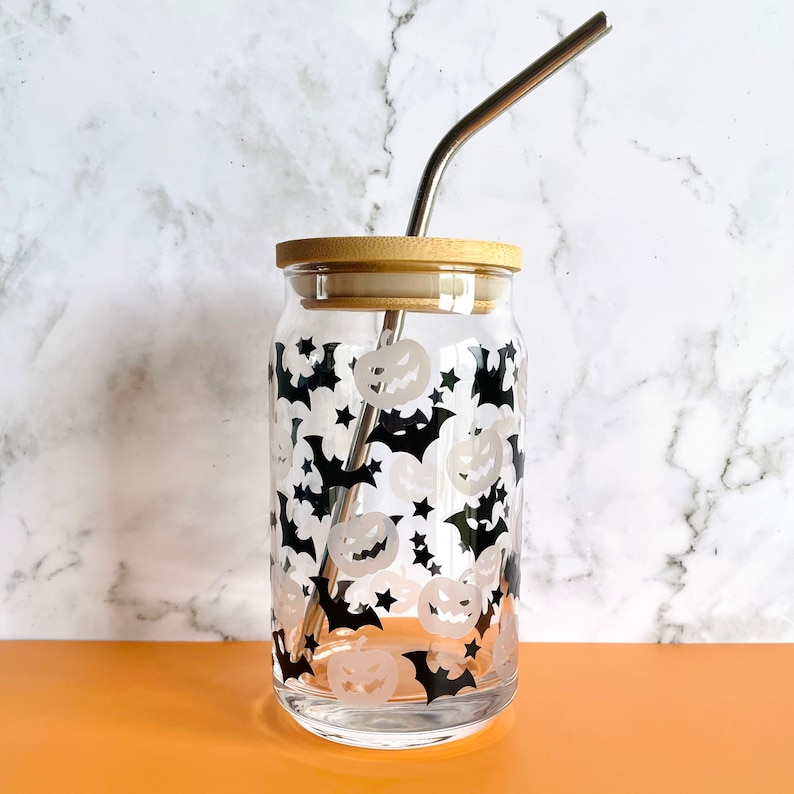 May include: A clear glass reusable drinking cup with a black and white Halloween design featuring bats and pumpkins. The cup has a bamboo lid and a stainless steel straw.